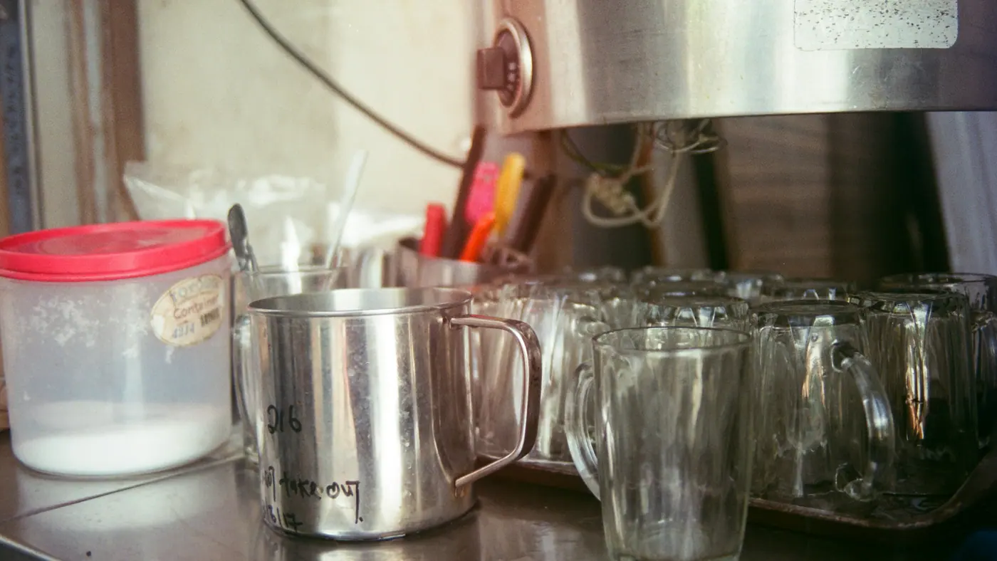 A kopitiam drinks counter in Singapore: steel cups, glasses and a jar of sugar on a tiled counter
