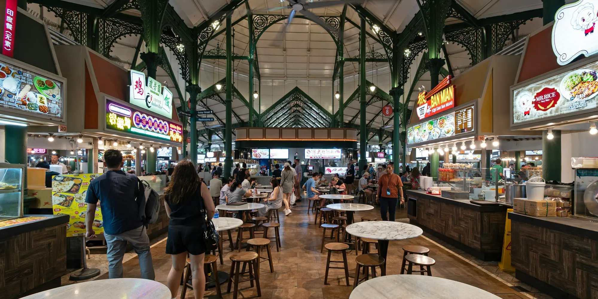 A hawker centre in Singapore under a cast-iron roof, people at round marble tables